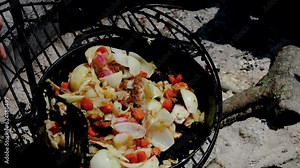 Using spatula to stir wild game meat being cooked over camp fire in cast iron skillet full of onions and carrots.