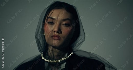 A serious fashionable woman looks at the camera posing in the studio, close-up of her face. A black veil on her face. The light turns on and off