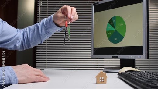 Person's hand displaying keys above a small wooden house while a computer monitor shows a financial pie chart representing property market data and investment analysis