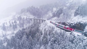 Download GRAUBUNDEN, SWITZERLAND - FEBRUARY 11, 2019 Viaduct and Glacier Express Train in Winter Day. Snowing. Swiss Alps. Switzerland. Aerial View. Drone Follows Train