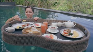 A beautiful asian girl in a swimming pool with a floating breakfast. Woman eating omlette. A girl is having breakfast in a swimming pool from a floating basket against the background of a green forest