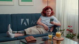 Attractive happy 35-40 disabled woman with artificial bionic prosthetic leg using smartphone to work online at cozy home