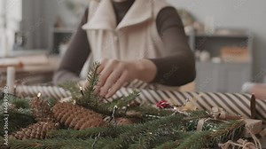Racking focus from Christmas tree branches on desk to blue-eyed Caucasian craftswoman decorating small gift box, then getting up and relocating to table
