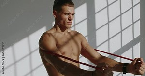 Focused young man with athletic body exercising with resistance band and training arm muscles at studio. Shirtless male has workout with sport equipment.