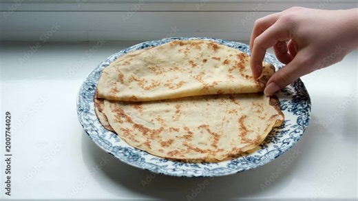 hand holding spoon over glass jar of red caviar next to large stack of golden crepes on white plate set on dark wooden table, concept of traditional russian cuisine, festive serving, homemade.