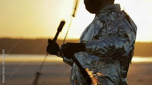 Medium shot of a pyrotechnic man in a silvery suit, who rotates torches on chains burning with fire with his hands as the sun sets in the middle of the desert.