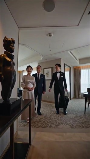 A bellboy guides a wealthy couple through a suite at a top-class hotel, showing them the beds. Th...