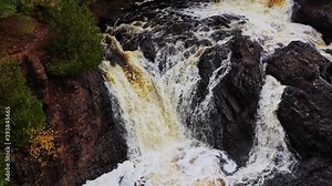 A close up aerial view of one of the waterfall cascades of Upper Potato River Falls as the heavy water flow tumbles down the red and brown carved rocky cliffs to the riverbed below.