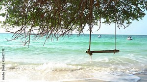 4K Landscape stabilize medium shot of empty wooden swing hanging under the tree on tropical sand beach. The sea waves rolling splash against the beach with floating jet ski and blue sky backgrounds.