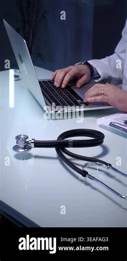 Medical doctor using a laptop and writing notes on a form, symbolizing modern healthcare data management, patient diagnosis, and professional medical consultation in an office with a stethoscope Stock Video Footage - Alamy