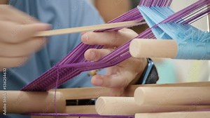 Hands and fingers using hand looming device to weave and adjust purple colored yarn to create traditional Asian belt, filmed as close up