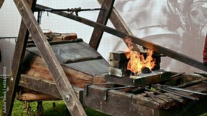 The blacksmith inflates the bellows of the brazier, and then with tongs removes a hot metal part from the fire.
