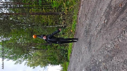 Vertical slow motion video of a man with black clothing throwing a Halloween pumpkin to pieces on the ground in a rural road with forest in the background. Pumpkin explodes to pieces after impact. Stock Video