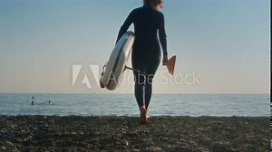 Senior woman on the beach in a wetsuit enters the water with a paddle board. An old female surfs at sunset at dusk. Active old age on the ocean. Granny steps into the sea with an oar in her hands.