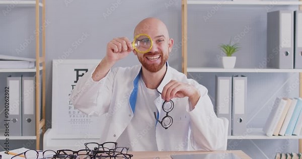 Young male medic ophthalmologist doctor at office desk with prescription glasses during optical eye checkup ophthalmic diopter visual consultation looks through loupe blinking with funny magnified eye