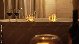 Wine and scented candles next to bathtub for romantic date night. Warm water filling up bathtub as candles burn in foreground with red wine and bottle. Hotel spa or cabin lodge. Close up