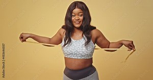 Unhappy black woman measuring body with tape. Upset African American plus size female in sportswear measuring chest and waist with tape on beige background in studio