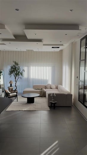 This living room feels expensive because the design focuses on light, space, and materials instead of decoration. The large curtains soften the daylight, making the room feel bright but calm. When light is filtered like this, every texture looks smoother and more premium. The ceiling has layered shapes with hidden lighting and small spotlights that add depth to the space. This makes the room feel more architectural and custom designed. High end interiors often use indirect lighting so the atmosp