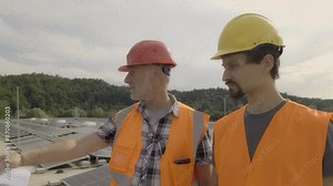 Electricians discussing solar panels array installation and maintenance on rooftop. Two male workers using digital tablet and draft while inspecting photovoltaic modules on roof, medium zoom in shot