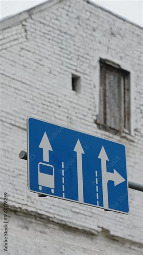 Blue traffic sign indicating lane directions with dedicated bus lane, two straight-ahead lanes, and right-turn-only lane, mounted on a pole against a white brick building facade.