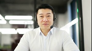 Webcam view. Asian corporate operator who works in customer support speaks on a video call looking in camera. Call center agent in wireless headset helping customer with complaints in business office