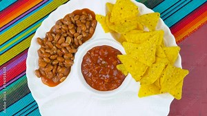Happy Cinco de Mayo party table with food platter including limes, corn chips, chilli beans and salsa on a red wood background.