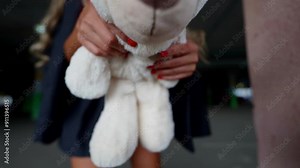 A beautiful woman with a teddy bear in her hands walks around the parking lot, wearing a black skirt and a pink top. Her outfit and joyful expression create a pleasant impression.