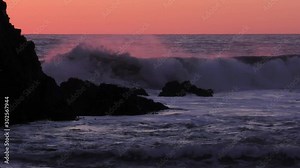 Amazing pink ocean waves crash against the dark blue rocks as the morning light skims the cresting sea water in a very complimentary colored artistic way.