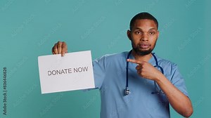 Portrait of male nurse holding donate now sign, inviting people to contribute, studio background. Healthcare worker in need of monetary support for patient care and medical research, camera A