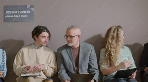 Tracking shot of diverse job candidates sitting in queue in office and discussing questions in application forms before interview