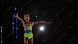 Slim cute attractive little girl athlete in bright colorful swimsuit performs elements of rhythmic gymnastics with hoop in dark gym with beautiful staged light and rear light
