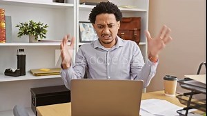 Frustrated young african american business worker losing it at the office, sitting mad and crazy at the table, shouting with arms raised in rage.