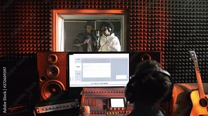Dolly-in shot of black man and woman dancing and singing into microphone in isolation booth of recording studio as male sound engineer using mixing desk in control room