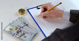 A cryptanalyst draws on paper a graph of the growth of the price of bitcoin on the exchange, calculating the dynamics of growth, next to it are American dollars and a stack of bitcoins.