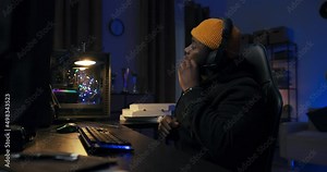 A professional programmer sits late into the night at a lit computer, writing software scripts, checking databases, doing remote infromatics work, talking to a co-worker through a headset.
