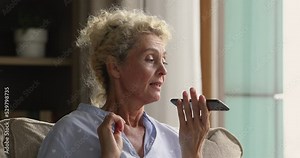 Focused senior mature lady talking on mobile phone call on speaker on mobile phone, speaking, listening, getting cheerful, smiling, laughing, using smartphone at home, recording audio message
