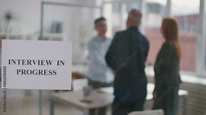 Interview in progress sign printed on paper hanging on glass wall in workspace, blurred people shaking hands and leaving in conference room on background