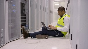 Stressed engineer sitting on floor and working on laptop, checking the servers in a data center.
