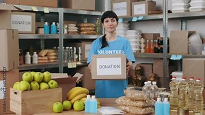 Portrsit of caucasian young woman standing at food bank warehouse and holding cardboard donation box. Charity organization and volunteering concept.