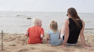 Woman with children sitting on the beach. Cute family looks at the waves in the sea on a hot summer day. Mom with her little daughter and son relaxing by the ocean