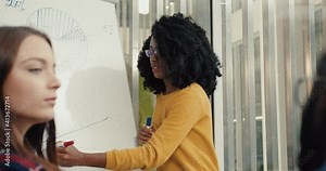 Young beautiful african american girl with glasses explains to group of students the topic of lecture and draws graphs on blackboard.