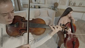 Top view of female string duo, young women playing cello and violin at minimalist luxury home interior. Closeup of musician practicing strings in light room, handheld shot