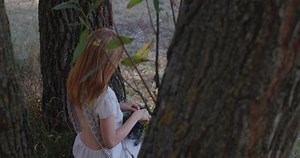 Beautiful blonde woman sitting under tree and weaving wreath of wild flowers on nature background. Portrait of a young girl with long hair in rustic linen dress and ethnic jewelry on sunny summer day.