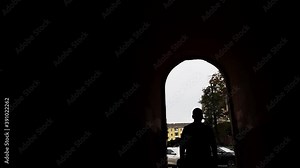 In the backlight, a man walks through a tunnel against a city background. A man walks into an oval arch in a residential building.