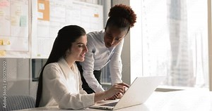 Young women colleagues working on joint on-line task completion, consider ideas. Female explain to co-worker new corporate software sit at desk look at laptop learn program. Apprenticeship, teamwork