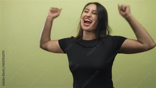 Woman cheering with open mouth and raised arms, young, wearing black top in green studio; excitement celebration.