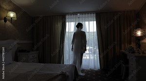 Bride in white boudoir dress stay near window and opening curtains. Wedding morning preparations. Luxury girl with fashion hairstyle in hotel room or apartment. Lovely woman in night gown and veil