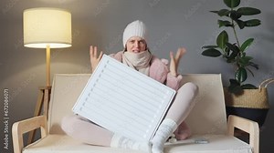 Angry woman wearing winter wool coat and hat sitting with electric oil heater and looks sadly, being frozen. exclaiming with negative facial expression.