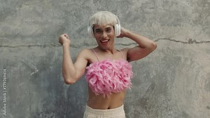 Portrait of the Cheerful Transgender Man Dancing in Pink Top Using Headphones. Handsome Positive Gender-Bending Person Listening to Music in the Middle of the Street and Dancing. LGBT People Concept