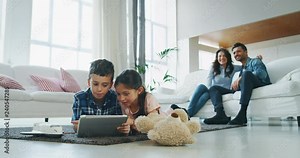Portrait of happy kids using tablet having fun lying on the carpet in living room with their parents in slow motion. Shot with RED camera in 8K. Concept of family entertainment, education, technology